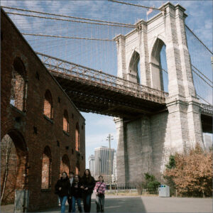Group of Sixth Formers walking on a street under the Brooklyn Bridge, with brick arches on the left and city buildings in the distance.