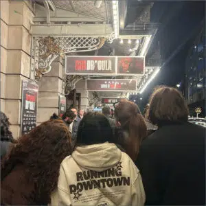 People line up outside a theater at night, passing under illuminated Dracula/Cynthia Erivo marquee and ornate ironwork signage.