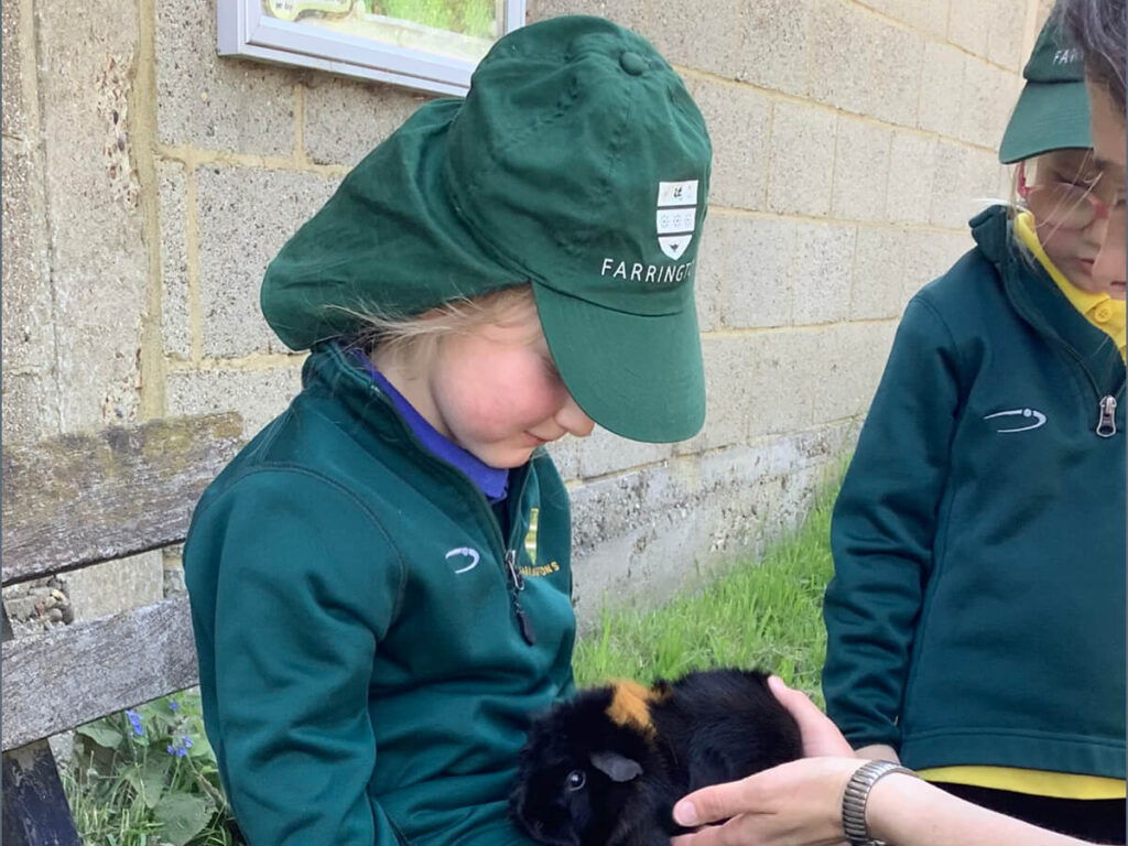 Two children in green uniforms sit outdoors, with one gently cradling a small black guinea pig with brown markings while an adult steadys the animal on her lap.