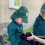 Two children in green uniforms sit outdoors, with one gently cradling a small black guinea pig with brown markings while an adult steadys the animal on her lap.
