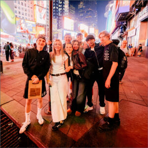 Group of Sixth Form students posing together at night in Times Square with bright billboards behind them, all smiling at the camera.