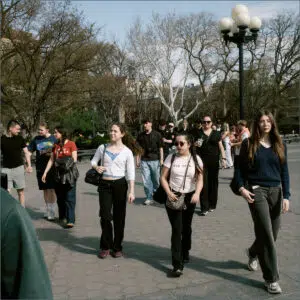 Group of Sixth Form students walking together in a sunny park plaza, with bare trees and a tall lamppost in the background, on a clear day.
