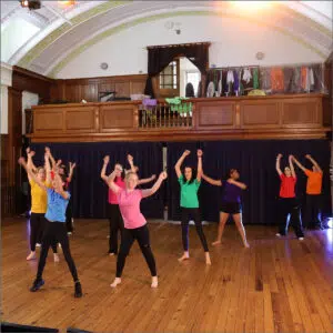 Group of children dancing on a wooden stage in colorful shirts, arms raised, with a balcony and dark curtain backdrop above them.