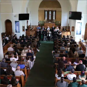 Choir performing at the front of a church nave with the audience seated in pews and some people filming with phones.