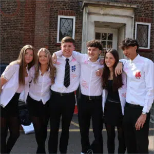 Six high school students in white shirts with pink signatures pose with arms around each other outside a brick building, smiling for a group photo.