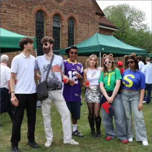 Six friends pose for a group photo at an outdoor campus event in front of a brick building and green tents, holding drinks and smiling.