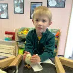 Young child gently holding a fluffy yellow chick in a classroom setup with toy shelves nearby.