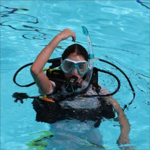 Person snorkeling in a bright blue pool, wearing a snorkeling mask and breathing tube with a buoyant vest, raising one arm above the water edge-to-edge view of the scene.