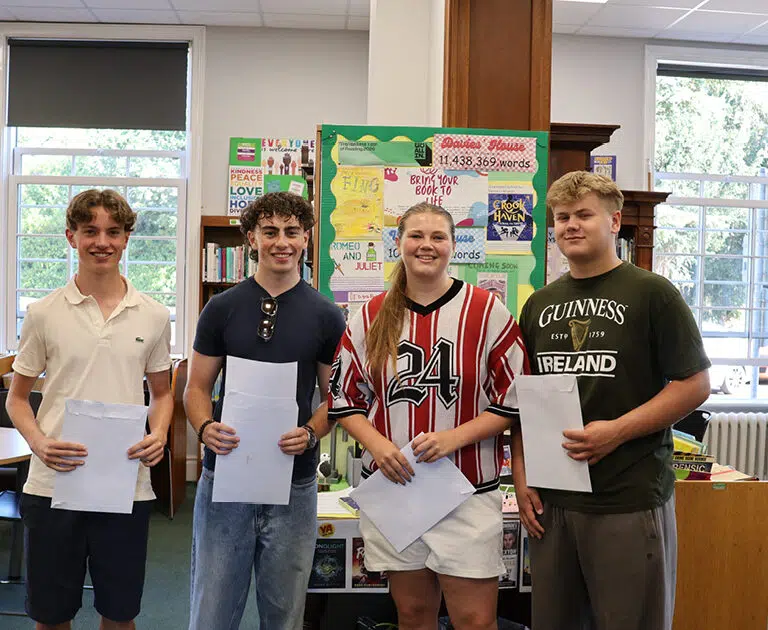 Four teens stand in a library, each holding a sheet of paper.